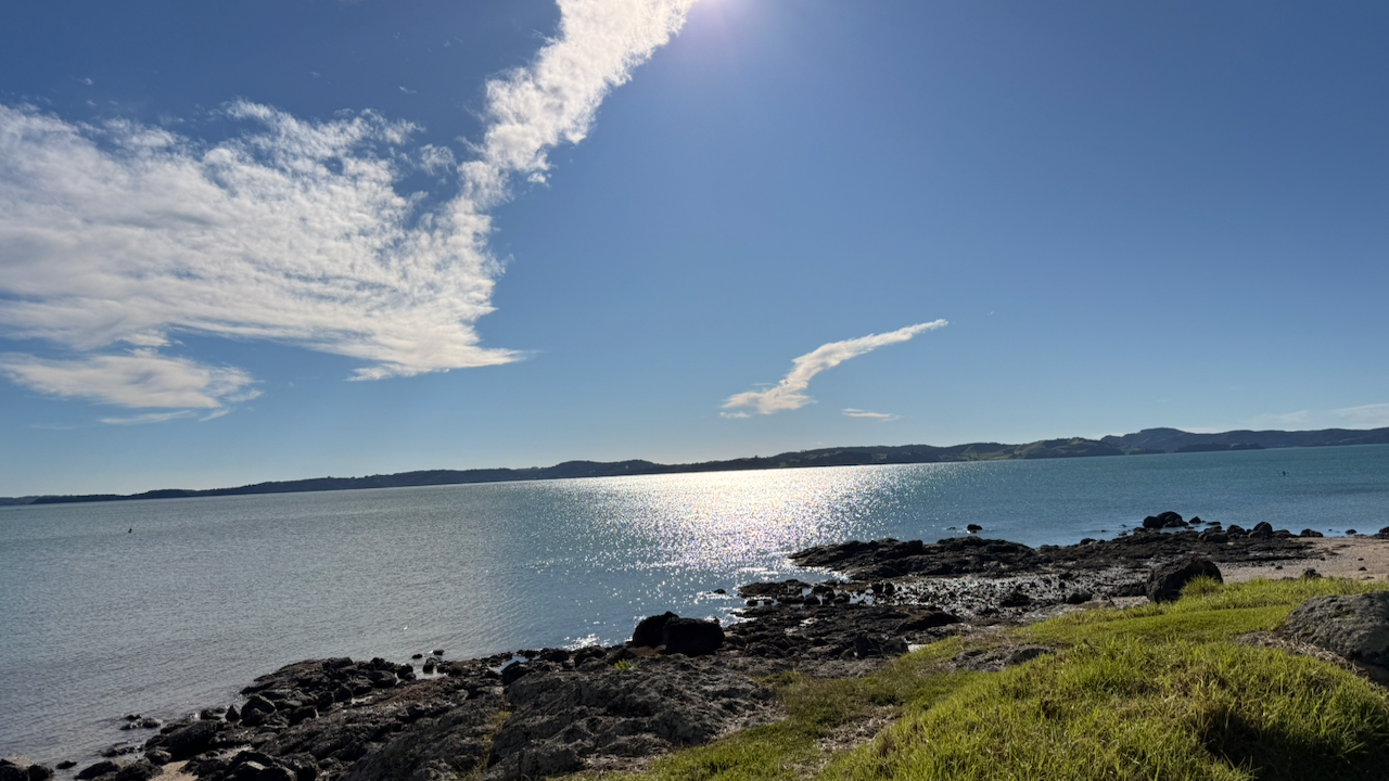 Rocky coastline with green grass, sun reflecting on calm harbour water under dramatic clouds